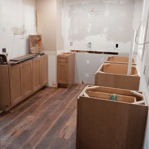 Early phase of kitchen remodel showing newly installed lower cabinets, unfinished walls, and wood-grain vinyl flooring preparation.